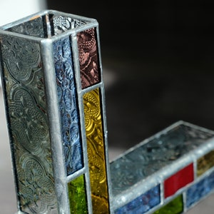 May include: A stained glass vase constructed with rectangular panels of textured glass in shades of clear, blue, yellow, green, and red, held together by silver-toned metal solder lines forming a geometric, L-shaped structure with an open top.