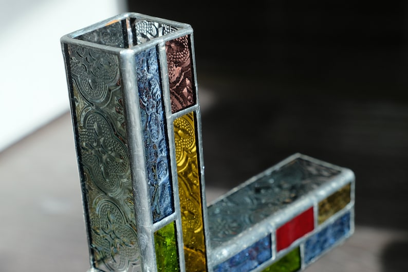May include: A stained glass vase constructed with rectangular panels of textured glass in shades of clear, blue, yellow, green, and red, held together by silver-toned metal solder lines forming a geometric, L-shaped structure with an open top.