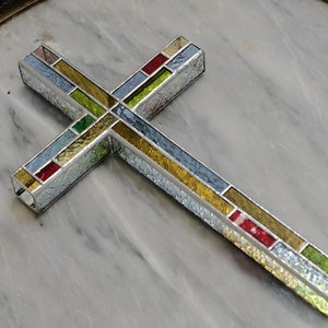 May include: A decorative cross constructed from multiple rectangular glass panels held together by metal framing. The glass features a variety of textures and colors, including clear, textured white, yellow, red, and green segments arranged in a grid.