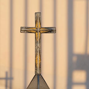 May include: A decorative cross featuring a metallic frame with a textured, amber-colored glass inlay. The cross is mounted on a triangular base constructed from thin, dark metal bars, creating a geometric structure with a warm, golden appearance.