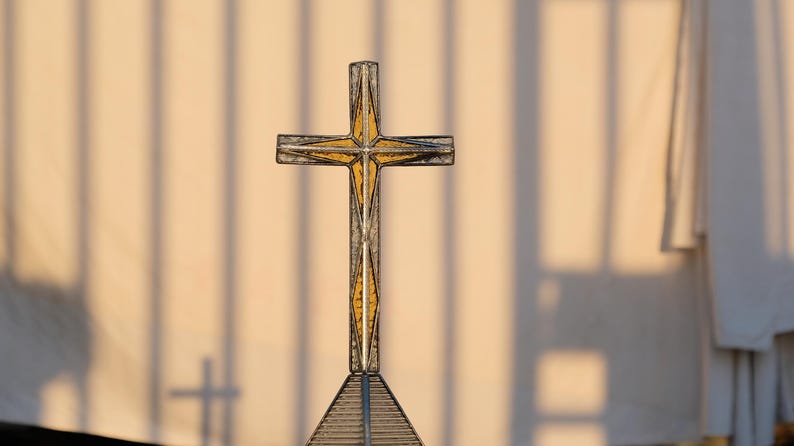 May include: A decorative cross featuring a metallic frame with a textured, amber-colored glass inlay. The cross is mounted on a triangular base constructed from thin, dark metal bars, creating a geometric structure with a warm, golden appearance.