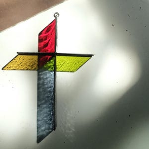 May include: A three-dimensional stained glass cross featuring a vertical segment with a red top and a textured gray bottom, intersected by a horizontal yellow textured glass bar. The cross is constructed with dark metal framing and a small hanging loop.