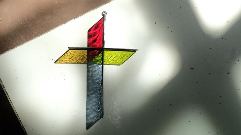 May include: A three-dimensional stained glass cross featuring a vertical segment with a red top and a textured gray bottom, intersected by a horizontal yellow textured glass bar. The cross is constructed with dark metal framing and a small hanging loop.