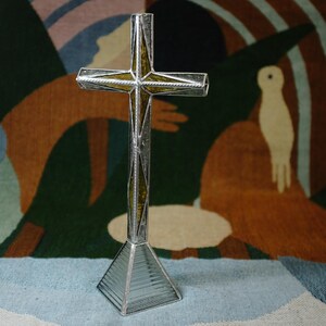 May include: A standing cross sculpture constructed from silver-toned metal framing and textured glass panels. The cross features a central vertical and horizontal bar with amber-colored glass inserts, supported by a clear, ribbed glass pyramid base.