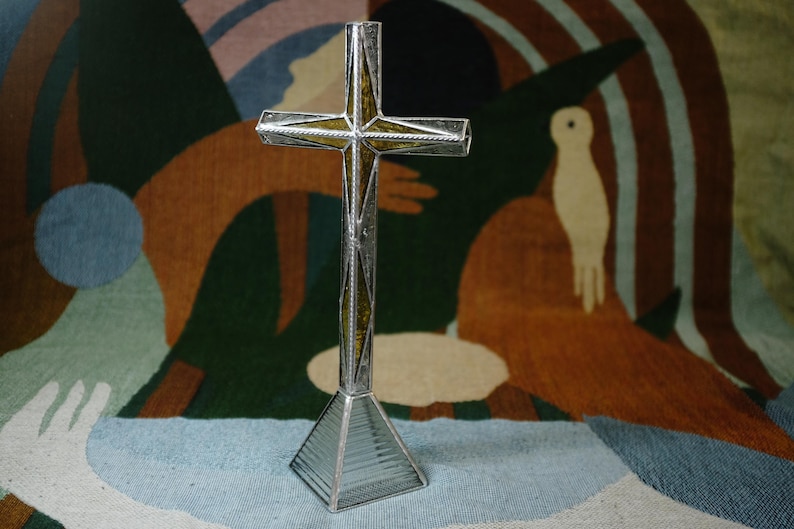 May include: A standing cross sculpture constructed from silver-toned metal framing and textured glass panels. The cross features a central vertical and horizontal bar with amber-colored glass inserts, supported by a clear, ribbed glass pyramid base.