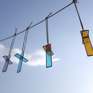 May include: Four stained glass cross ornaments hanging from a horizontal string. The crosses feature various colors including purple, light blue, red, green, and yellow, all framed in dark metal solder lines against a bright blue sky background.
