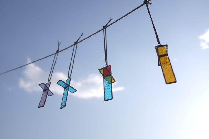 May include: Four stained glass cross ornaments hanging from a horizontal string. The crosses feature various colors including purple, light blue, red, green, and yellow, all framed in dark metal solder lines against a bright blue sky background.