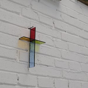 May include: A wall-mounted cross constructed from four distinct pieces of stained glass. The top section is red, the horizontal arms are yellow and green, and the bottom vertical section is blue. Each piece is joined by thin black metal framing.