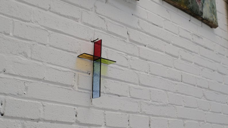 May include: A wall-mounted cross constructed from four distinct pieces of stained glass. The top section is red, the horizontal arms are yellow and green, and the bottom vertical section is blue. Each piece is joined by thin black metal framing.