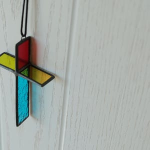 May include: A three-dimensional stained glass cross ornament featuring intersecting panels of red, yellow, and blue textured glass held together by dark metal framing. The cross hangs from a thin black cord, displaying vibrant, translucent colors.