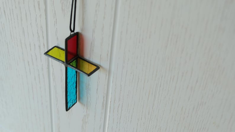 May include: A three-dimensional stained glass cross ornament featuring intersecting panels of red, yellow, and blue textured glass held together by dark metal framing. The cross hangs from a thin black cord, displaying vibrant, translucent colors.
