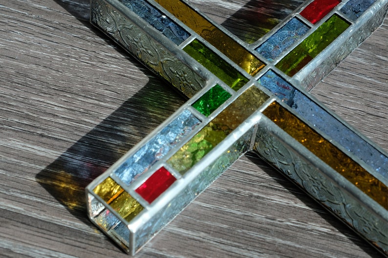 May include: A three-dimensional cross constructed from metal frames and multi-colored stained glass panels. The glass features shades of red, yellow, green, and blue, with some sections displaying a textured, iridescent finish and embossed metal sides.