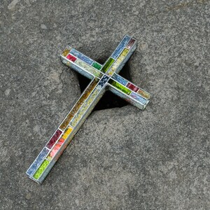 May include: A cross-shaped object constructed with a silver-toned metal frame and filled with small, multi-colored glass mosaic tiles in shades of red, yellow, green, and blue, arranged in a grid pattern along the length of the cross arms.