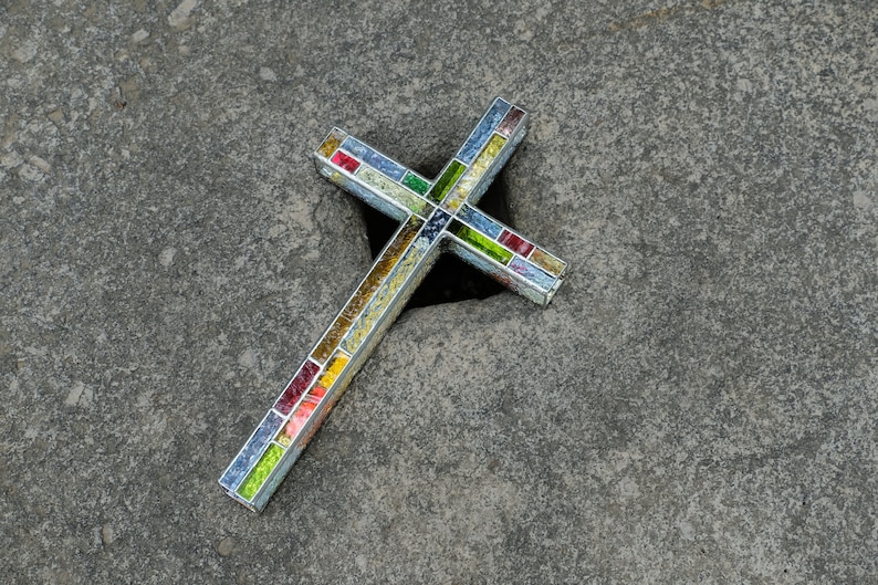 May include: A cross-shaped object constructed with a silver-toned metal frame and filled with small, multi-colored glass mosaic tiles in shades of red, yellow, green, and blue, arranged in a grid pattern along the length of the cross arms.