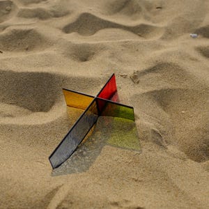 May include: A cross-shaped sculpture constructed from four intersecting translucent acrylic panels in distinct colors, including red, yellow, green, and dark blue, casting a sharp, colorful shadow onto the surrounding textured sand surface.
