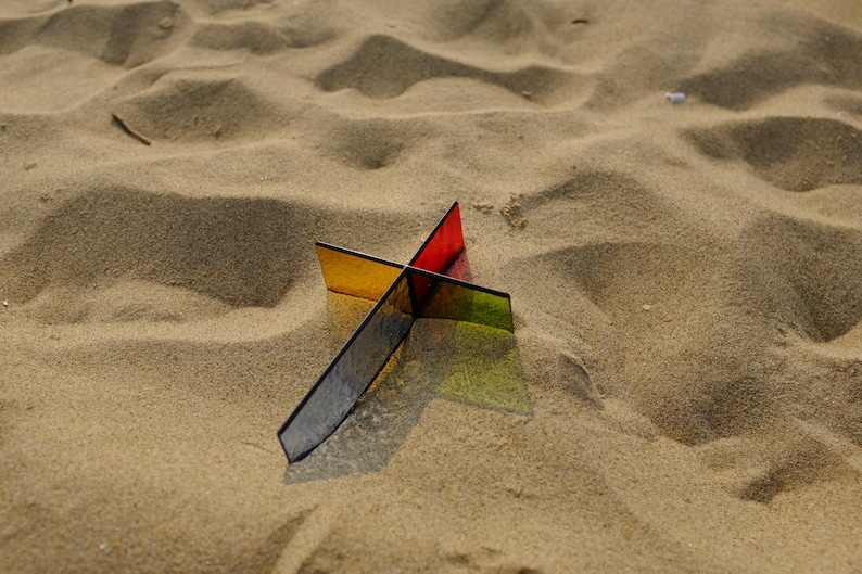 May include: A cross-shaped sculpture constructed from four intersecting translucent acrylic panels in distinct colors, including red, yellow, green, and dark blue, casting a sharp, colorful shadow onto the surrounding textured sand surface.