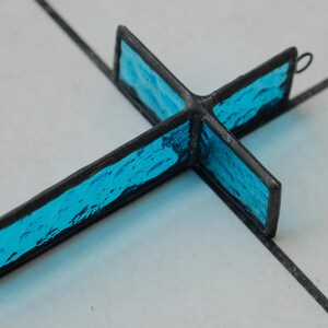 May include: A cross-shaped ornament constructed from textured, vibrant blue glass panels encased in dark metal soldering. The piece features a small circular metal loop at the top edge for hanging, showcasing a translucent, rippled glass surface.