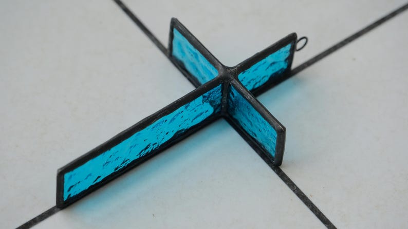 May include: A cross-shaped ornament constructed from textured, vibrant blue glass panels encased in dark metal soldering. The piece features a small circular metal loop at the top edge for hanging, showcasing a translucent, rippled glass surface.