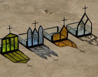 May include: Four small, open-topped containers constructed from stained glass and dark metal frames. Each piece features a cross at the peak, with panels in lime green, textured clear, amber, and clear glass, casting distinct shadows on the ground.