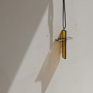May include: A cross pendant made of translucent yellow glass with a dark metal frame, suspended from a thin black cord. The cross features a textured surface and is positioned to cast a distinct, elongated shadow on the wall behind it.