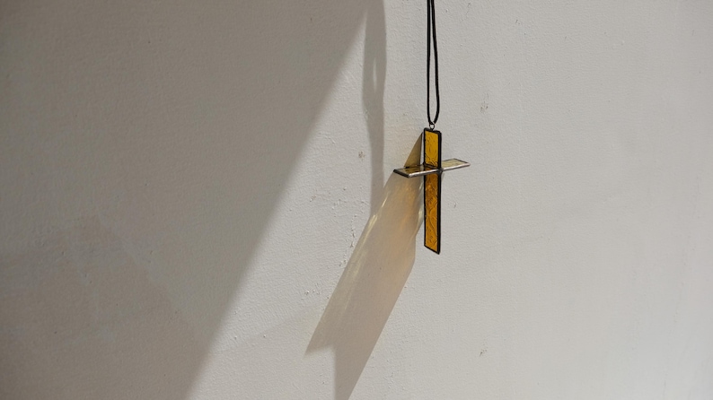 May include: A cross pendant made of translucent yellow glass with a dark metal frame, suspended from a thin black cord. The cross features a textured surface and is positioned to cast a distinct, elongated shadow on the wall behind it.