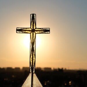 May include: A textured glass cross with a dark metal frame, positioned vertically against a bright, glowing sunset. The light shines through the translucent material of the cross, highlighting its geometric structure and clear, crystalline surface.