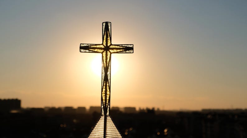 May include: A textured glass cross with a dark metal frame, positioned vertically against a bright, glowing sunset. The light shines through the translucent material of the cross, highlighting its geometric structure and clear, crystalline surface.