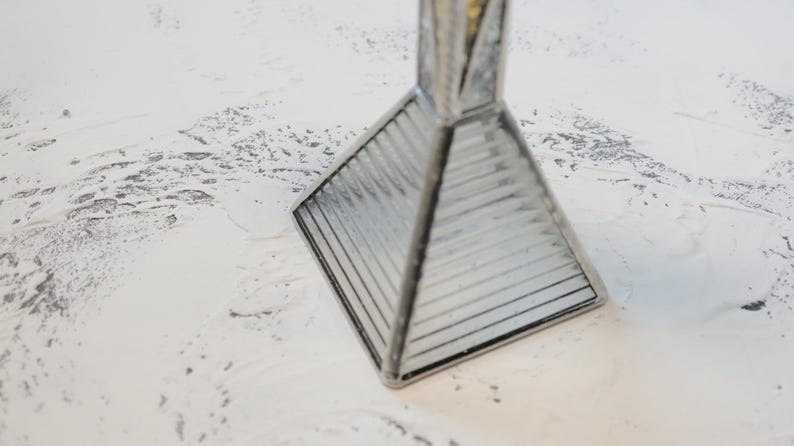 May include: A pyramid-shaped glass object with a square base and ribbed, clear glass panels held together by dark metal soldering. The structure tapers to a point at the top, featuring a reflective, geometric design with clean, straight lines.
