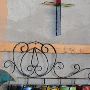 May include: A wall-mounted black metal rack with decorative scrollwork holds four painted metal cans in blue, yellow, green, and red. Above the rack, a stained glass cross features a vertical black textured panel and a horizontal red panel.