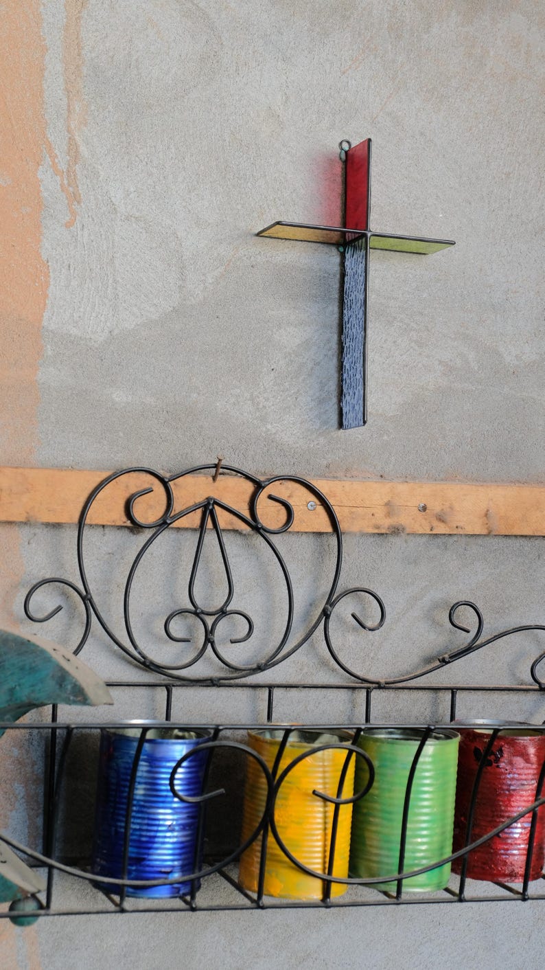 May include: A wall-mounted black metal rack with decorative scrollwork holds four painted metal cans in blue, yellow, green, and red. Above the rack, a stained glass cross features a vertical black textured panel and a horizontal red panel.