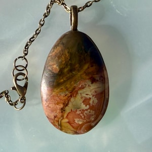 May include: Oval pendant necklace with a natural stone pendant. The pendant features a mix of colors, including brown, orange, and blue. The chain and pendant hardware are a bronze color. The necklace is a jewelry item.