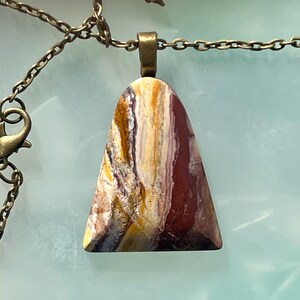 May include: A pendant necklace with a triangular stone in shades of red, yellow, and white. The stone hangs from a bronze-coloured chain with a matching clasp. The pendant displays a distinctive, natural pattern, perfect for everyday wear.