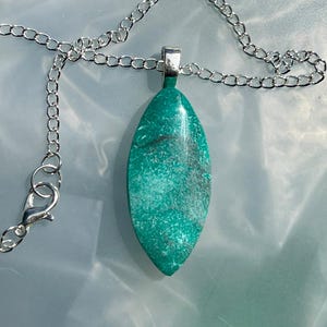 Puede incluir: Un collar de cadena de plata con un colgante de piedra preciosa verde. El colgante tiene forma de lágrima y presenta un patrón de mármol.