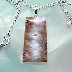May include: A rectangular pendant necklace with a silver chain. The pendant features a polished stone with a mix of brown, white, and red hues. The stone has a unique pattern with a central white area.