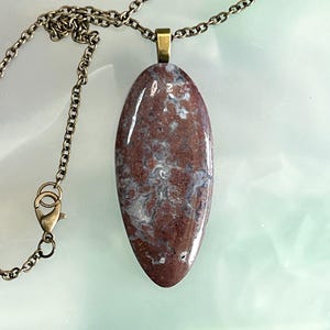 Puede incluir: Un collar con colgante que presenta una piedra ovalada pulida con patrones rojos, marrones y blancos. La piedra cuelga de una cadena de color bronce con un cierre a juego. El collar es una joya.