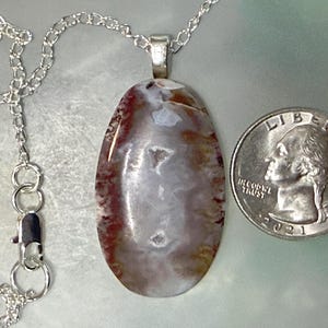 May include: An oval-shaped pendant featuring a polished stone with swirling patterns of white, grey, and reddish-brown, suspended from a silver-colored chain. A 2021 United States quarter coin is placed next to the pendant for size reference.