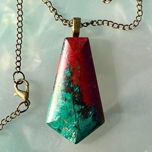 May include: A pendant necklace featuring a geometric, trapezoid-shaped stone. The stone displays a vibrant color palette of red, green, and black. The necklace has a bronze-colored chain and clasp, creating a striking contrast with the stone's colors.