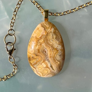 Può includere: Una collana color oro con un grande pendente a forma di goccia. Il pendente è una pietra lucida con motivi intricati nei toni del beige, marrone e crema. La collana ha una chiusura a moschettone.
