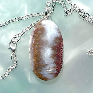 May include: A silver chain necklace with an oval pendant. The pendant features a natural stone with a mix of white, brown, and red hues. The chain is a classic link style with a lobster clasp.