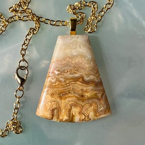 Può includere: Collana con catena color oro e pendente. Il pendente, di forma trapezoidale, presenta un motivo a strati di tonalità beige, marrone e marrone. La collana ha una chiusura a moschettone.