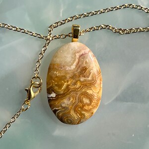 Op de afbeelding: Een goudkleurige ketting met een ovale hanger. De hanger is een gepolijste steen met een wervelend patroon van beige, bruin en wit. De ketting heeft een karabijnsluiting. De hanger is ongeveer 4 cm lang.