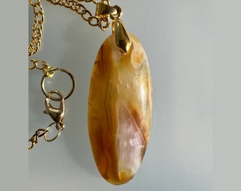May include: A gold-colored chain necklace with a large, oval pendant. The pendant is a mix of amber, cream, and brown tones, with a polished, smooth surface. The necklace has a lobster clasp closure.