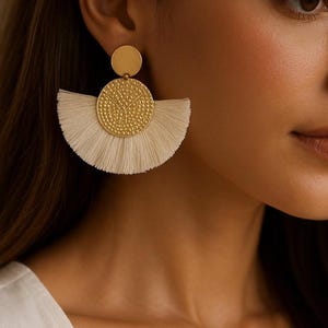 Peut inclure: Boucles d'oreilles pendantes dorées. Elles présentent un cercle doré sur le dessus et un pendentif en forme d'éventail texturé avec des franges crème. Ces boucles d'oreilles sont conçues pour ajouter de l'élégance.