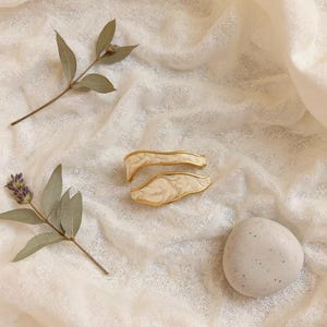 Peut inclure: Deux pinces à cheveux dorées avec un motif marbré blanc et crème. Les pinces sont posées sur un tissu crème, avec des brins de lavande et une pierre lisse et tachetée. Les pinces ont une forme ondulée.