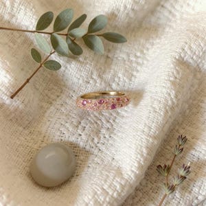 Peut inclure: Une bague en or ornée de petites pierres roses, posée sur un tissu blanc texturé. L'image comprend une branche de feuilles et une pierre grise lisse. Le design de la bague est délicat et féminin, idéal pour un cadeau.
