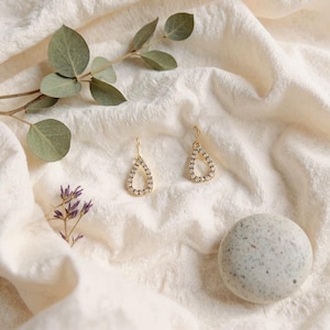 Peut inclure: Deux boucles d'oreilles dorées en forme de goutte d'eau, ornées de pierres claires et scintillantes. Elles sont posées sur un tissu crème texturé, avec une branche d'eucalyptus et une petite pierre tachetée.
