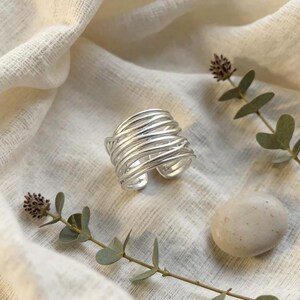Peut inclure: Une large bague en argent composée de plusieurs couches de fines bandes métalliques superposées qui s'enroulent autour du doigt. La bague présente une finition polie et une structure ouverte, révélant une série de boucles entrelacées.