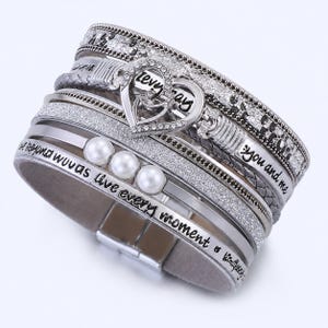 Op de afbeelding: Zilveren meerlaagse armband met een hartvormige bedel. De armband heeft imitatieparels, strass-steentjes en gegraveerde tekst met de tekst "love", "you and me" en "live every moment". De armband heeft een magnetische sluiting.