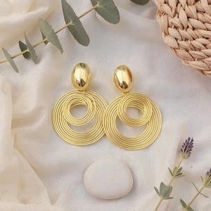 Peut inclure: Une paire de boucles d'oreilles dorées avec un motif en spirale. Chaque boucle d'oreille présente un grand pendentif en spirale circulaire et un petit clou de forme ovale. Les boucles d'oreilles sont présentées sur une surface blanche.