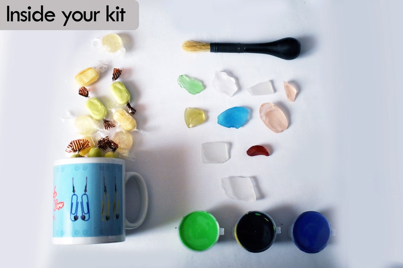 Puede incluir: Un kit de manualidades con una taza azul llena de caramelos, un pincel, vidrio de mar de varios colores y tres peque&ntilde;os botes de pintura en verde, negro y azul.